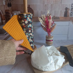 Main déroulant un rouleau essuie-tout lavable Muguet dans une cuisine avec rouleau à côté d'un camembert posé sur une planche et un bouquet de fleurs séchées