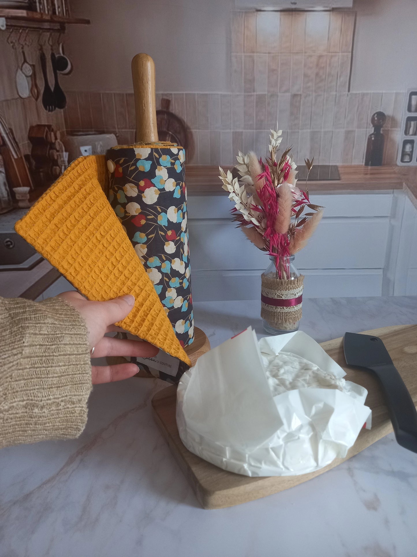 Main déroulant un rouleau essuie-tout lavable Muguet dans une cuisine avec rouleau à côté d'un camembert posé sur une planche et un bouquet de fleurs séchées