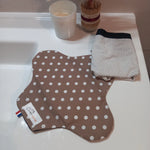 La serviette menstruelle lavable à pois blancs sur fond taupe est posée ouverte sur un rebord de lavabo, à côté d'un sous-vêtement plié. Le côté motif en coton est visible.