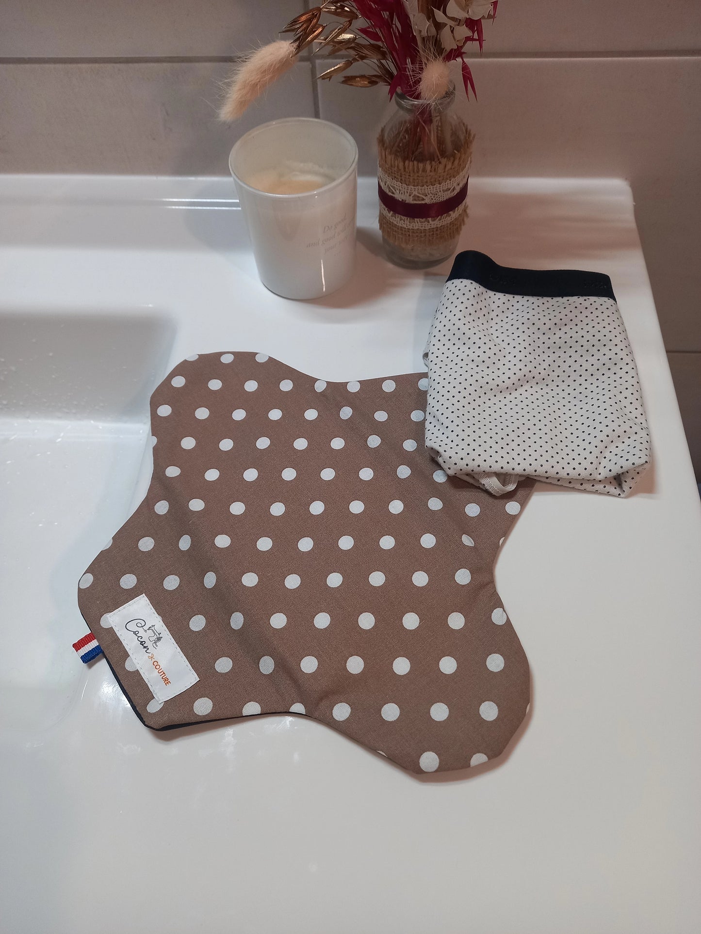 La serviette menstruelle lavable à pois blancs sur fond taupe est posée ouverte sur un rebord de lavabo, à côté d'un sous-vêtement plié. Le côté motif en coton est visible.