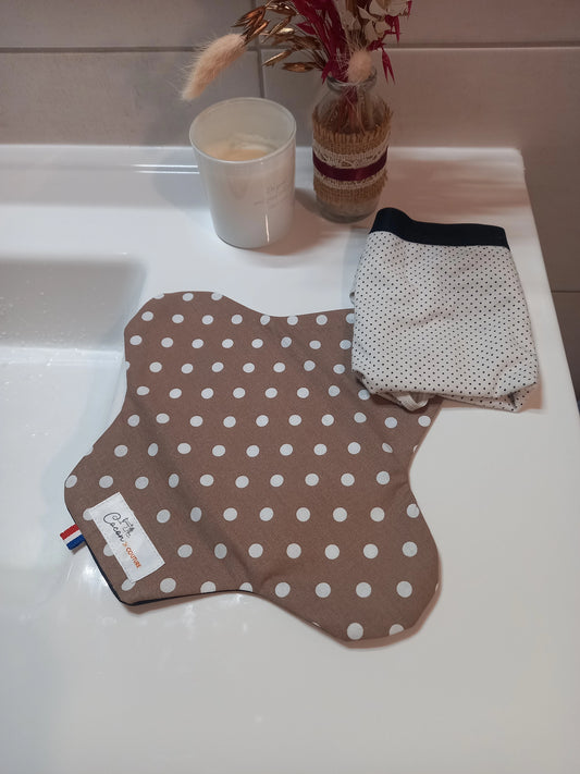 La serviette menstruelle lavable à pois blancs sur fond taupe est posée ouverte sur un rebord de lavabo, à côté d'un sous-vêtement plié. Le côté motif en coton est visible.