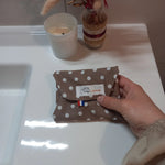 La serviette menstruelle lavable à pois blancs sur fond taupe est pliée et tenue dans une main. L'étiquette Cocon Couture et un drapeau français sont visibles sur le rabat.