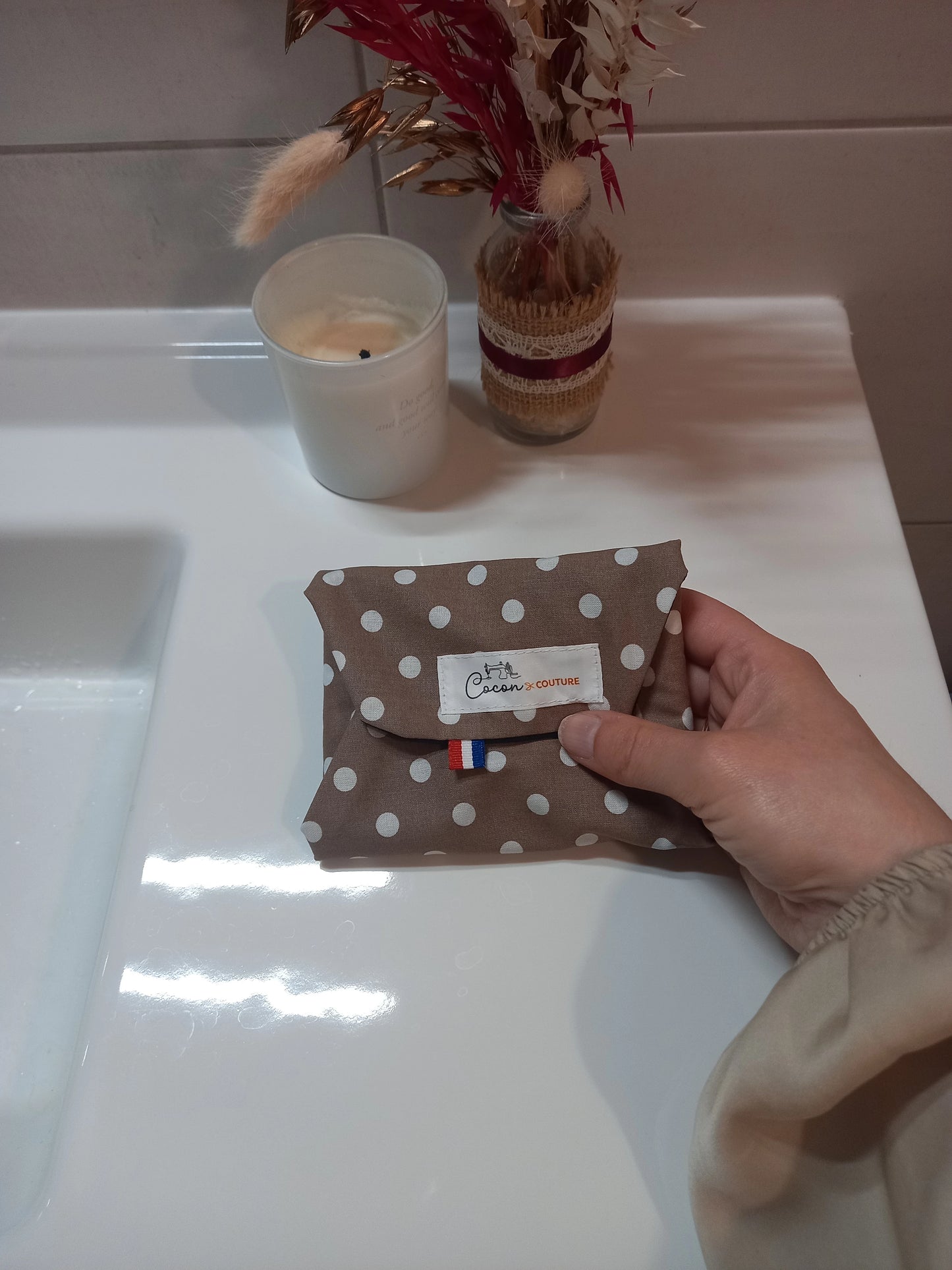 La serviette menstruelle lavable à pois blancs sur fond taupe est pliée et tenue dans une main. L'étiquette Cocon Couture et un drapeau français sont visibles sur le rabat.